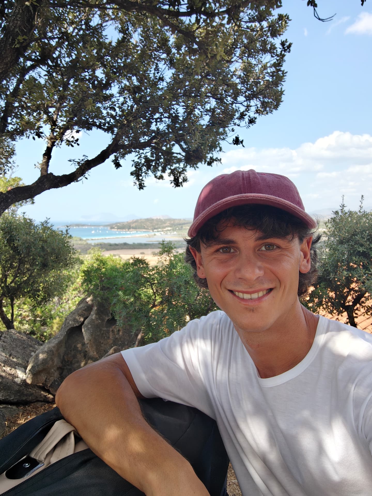 Simone sorride sotto gli alberi, con una baia e alcune barche all'ancora sullo sfondo